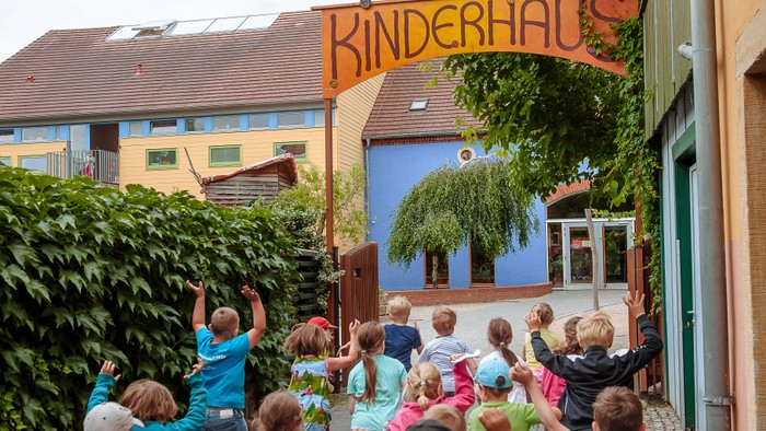 Kinder versammeln sich vor einem bunten Kinderhaus-Kindertagesstätte und winken begeistert, während im Hintergrund Bäume und Gebäude zu sehen sind.