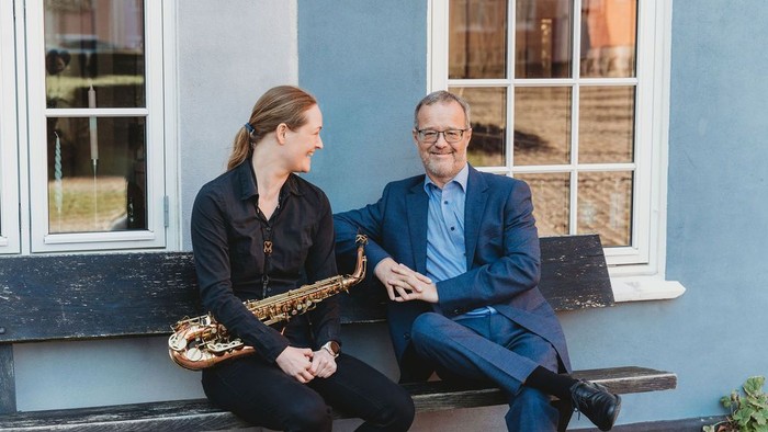 To personer sidder på en bænk, den ene holder en saxofon, og de smiler sammen udendørs.