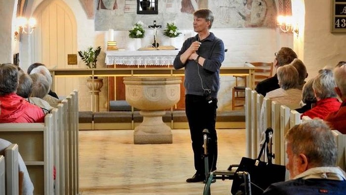 En mand står og tale i en kirke, mens en kamera holdes op på en tripod.