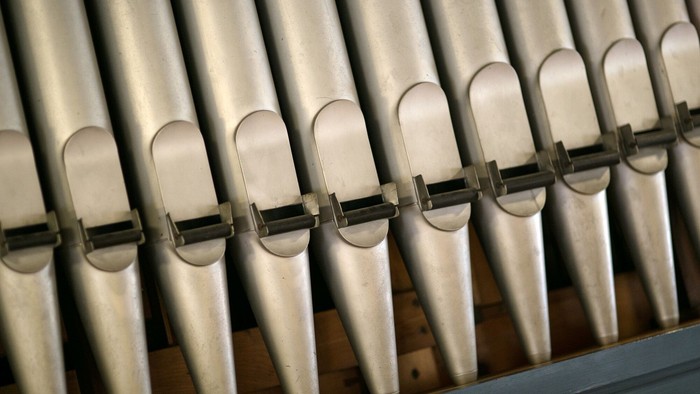Die Nahaufnahme zeigt mehrere Orgelpfeifen aus Metall, die in gleichmäßiger Reihe nebeneinander angeordnet sind. Die Pfeifen variieren leicht in Größe und Form, ihre oberen Öffnungen sind abgerundet. Eine Querstrebe mit kleinen Halterungen verläuft vor d