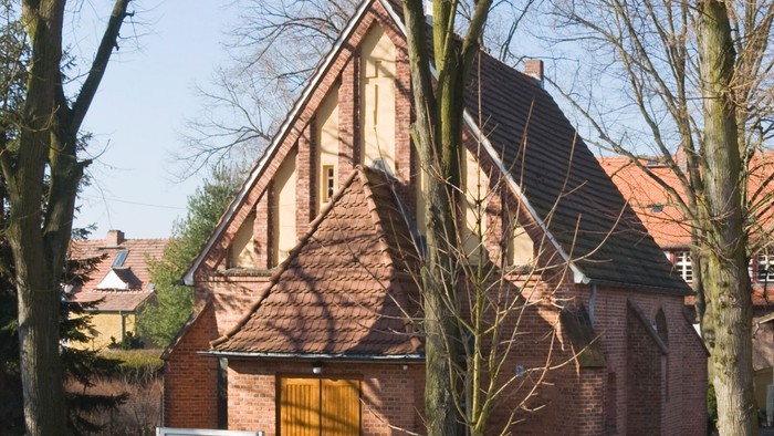 Historische Backsteinkirche mit steilem Satteldach und Holztür, umgeben von kahlen Bäumen.