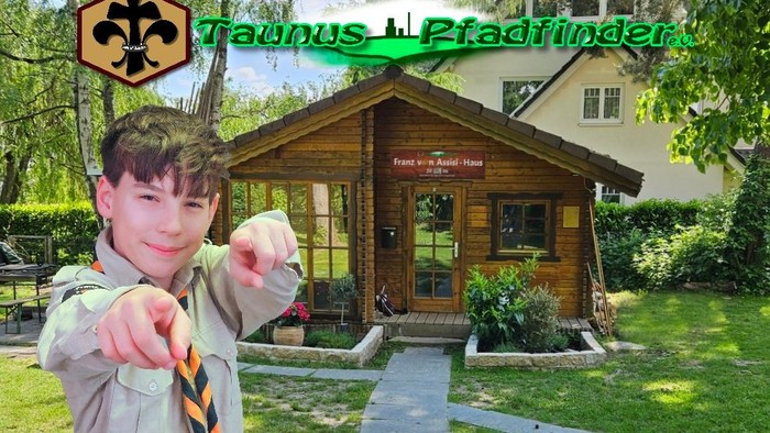 Junge in Pfadfinderuniform zeigt mit fröhlichem Gesichtsausdruck auf eine Holzhütte mit der Aufschrift „Taunus Pfadfinder“.