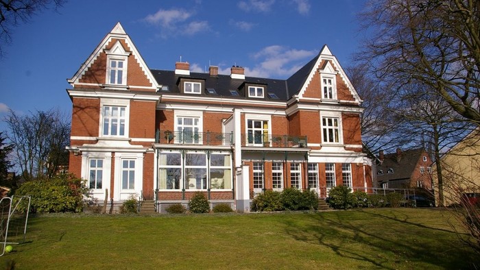 Großes zweigeschossiges Backsteinhaus mit weißen Zierleisten und zahlreichen Fenstern, das auf einem grasbewachsenen Rasen unter blauem Himmel steht.