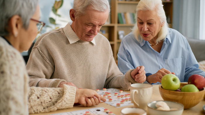 Drei ältere Menschen spielen ein Brettspiel, während frisches Obst und Tee auf dem Tisch stehen.