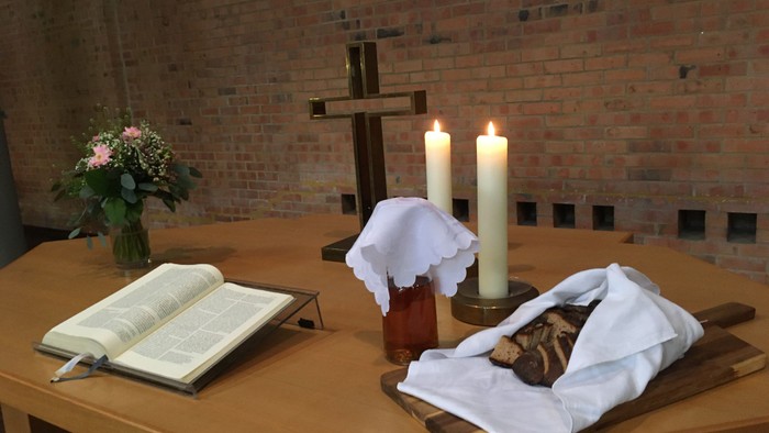 Religiöser Altaraufbau mit Kerzen, einem Kreuz, einem Buch, Blumen und einem bedeckten Laib Brot auf einem Holzbrett.