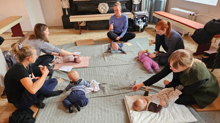 Gruppe af voksne og babyer deltager i en interaktiv gulvsession, sandsynligvis et forældre- eller spædbarnsplejekursus.