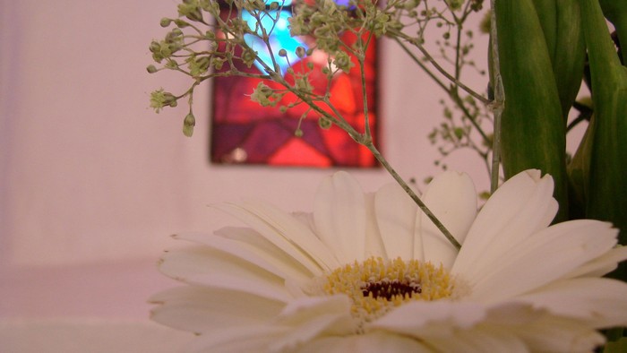 En blomstest med hvide gerberier og grønne blomster foran et billede med farverige glasmøbler.