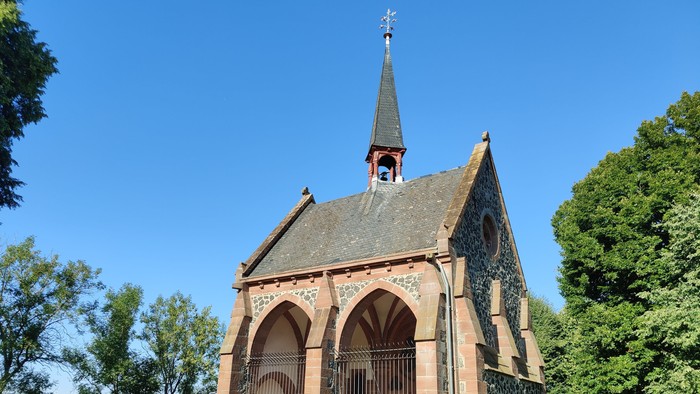 Idyllische Kapelle mit gewölbtem Eingang steht in einem ruhigen, gepflegten Friedhof unter klarem blauem Himmel.