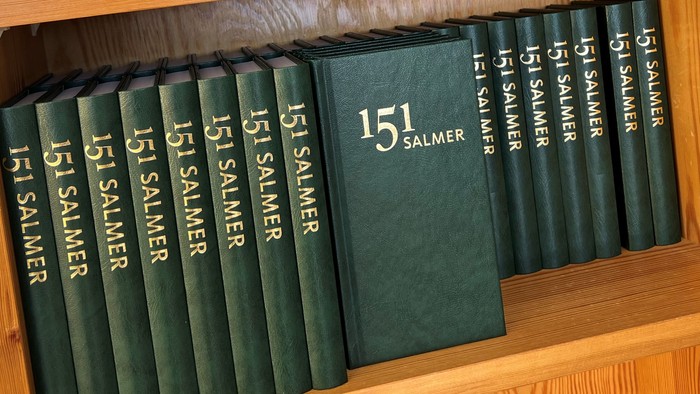 En samling af grønne 151 Salmer salmebøger pænt arrangeret på træhylder.