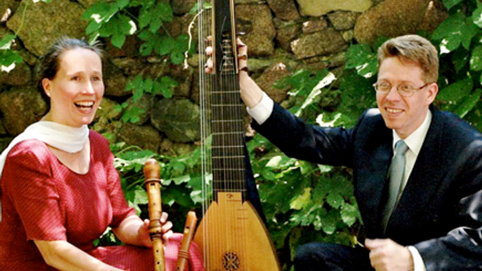 Zwei Personen spielen im Freien traditionelle Instrumente in der Nähe einer Steinmauer und lächeln sich an.