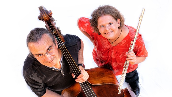 Zwei Musiker posieren gemeinsam, einer hält ein Cello und der andere eine Flöte, beide lächeln glücklich.