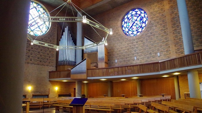 Moderne Kircheninnenausstattung mit hölzernen Kirchenbänken, einer Orgel und runden Buntglasfenstern mit warmem Licht.