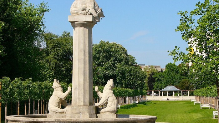 Parkbrunnen mit Statuen und Vögeln, umgeben von Grünflächen.