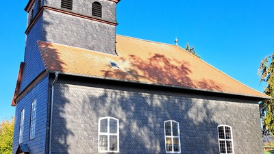 Historische Kirche mit hohem Kirchturm und weißem Kreuz vor einem klaren blauen Himmel.