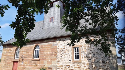 Steinerne Kirche mit roter Tür und Turmspitze, teilweise von Bäumen verdeckt.