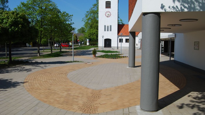 Ein moderner Platz mit einem weißen Uhrturm und einem gewundenen Sandweg.