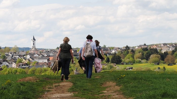 Mehrere Personen gehen auf einem schmalen Erdweg durch eine grüne Wiesenlandschaft. Sie sind von hinten zu sehen und tragen Rucksäcke oder Jacken über dem Arm. Der Weg führt leicht bergab in Richtung eines Ortes mit dicht stehenden Häusern. In der Mitte d