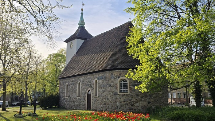 Kleine Kirche mit spitzem Turm, umgeben von Bäumen und blühenden Blumen