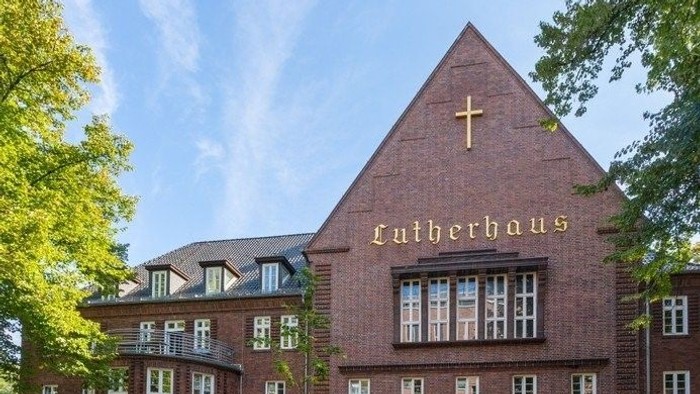 Backsteinkirche mit Kreuz und „Lutherhaus“-Beschilderung, umgeben von Bäumen.
