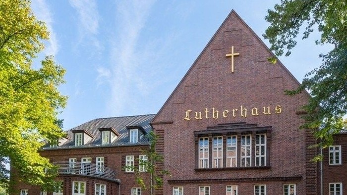 Backsteinkirche mit Kreuz und „Lutherhaus“-Beschilderung, umgeben von Bäumen.