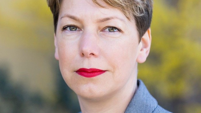 Professionelle Frau in einem grauen Blazer mit rotem Lippenstift, vor einem unscharfen Außenhintergrund.