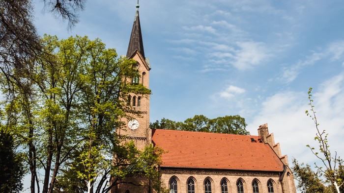Kirche mit hohem Turm und Uhr, umgeben von Bäumen unter blauem Himmel.