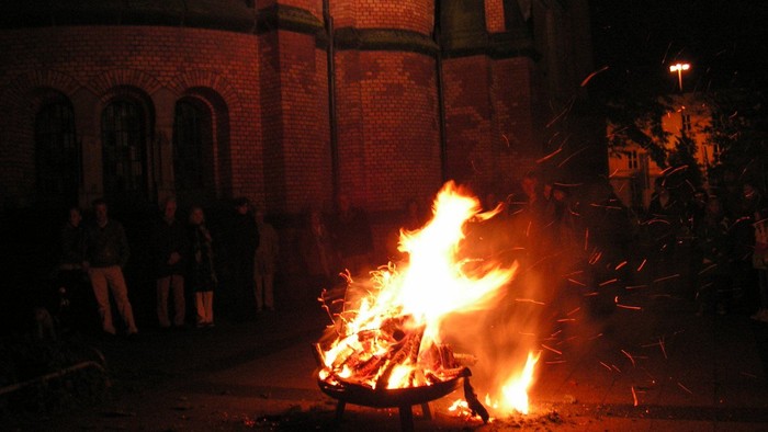 Ein nächtliches Lagerfeuer brennt hell, während sich im Hintergrund Schaulustige nahe einem Backsteingebäude versammeln.