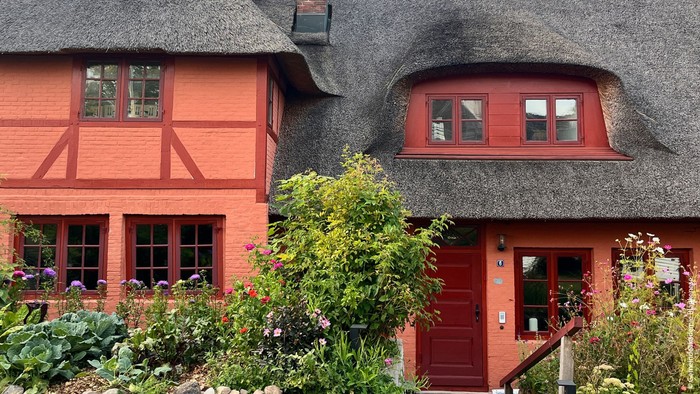 Bezauberndes Fachwerkhaus mit Reetdach und leuchtend roter Tür, umgeben von üppigem Grün und Blumen.