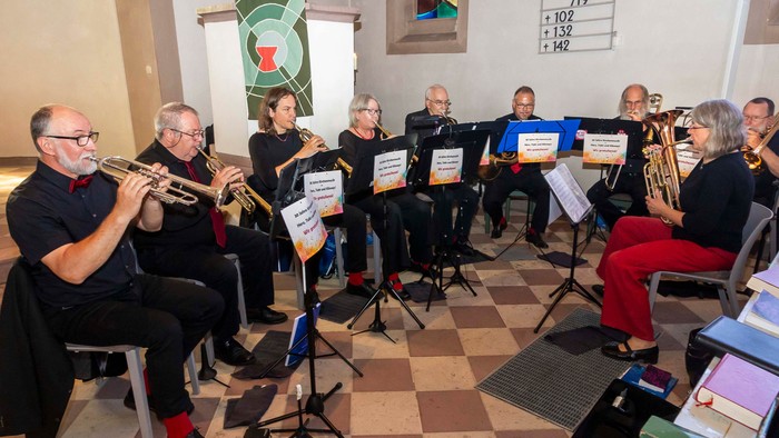 Eine Gruppe von Musikerinnen und Musikern sitzt im Halbkreis in einer Kirche und spielt Blechblasinstrumente. Zu sehen sind unter anderem Trompeten und Posaunen. Die Mitwirkenden sind schwarz gekleidet, einige mit roten Akzenten wie Fliegen oder Socken. 