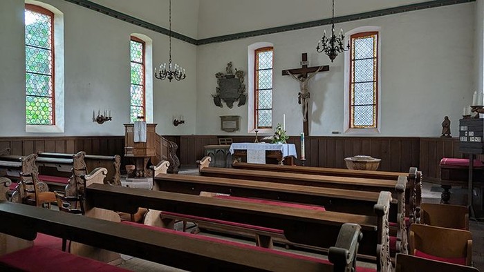 Das Bild zeigt den Innenraum einer kleinen, schlichten Kirche. Holzbänke mit roten Polstern sind in Reihen angeordnet und richten sich auf den Altarbereich aus. Vorne steht ein Altar mit weißer Decke, dahinter ist ein Kruzifix an der Wand angebracht. Meh