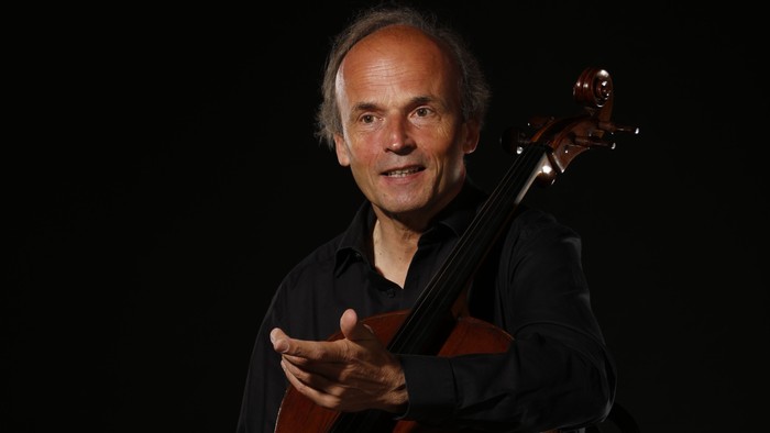 Ein Musiker spielt lächelnd Cello, während er in einer schwach beleuchteten Umgebung sitzt.