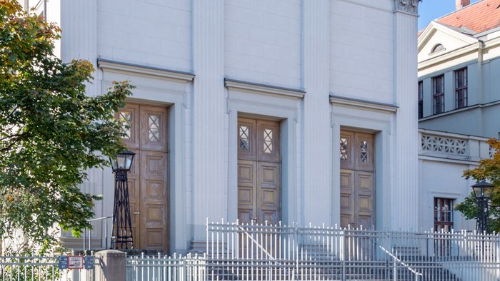 Das Bild zeigt eine weiße Kirche mit Säulen und einem Zaun davor, vor einem klaren blauen Himmel.