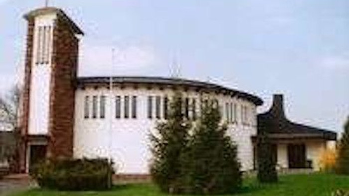 Das Bild zeigt eine Kirche mit einem hohen Turm und einem runden Anbau, umgeben von Grünflächen und Bäumen.