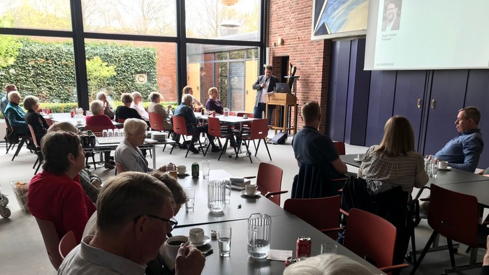 En gruppe af mennesker deltager i et møde i et rum med store vinduer og en projektor.