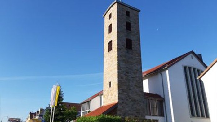 Moderne Kirche mit hohem Steinglockenturm und rotem Ziegeldach unter klarem blauem Himmel.