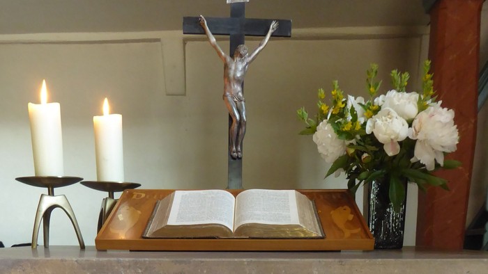 Eleganter Kirchenaltar mit Kerzen, einem Kreuz, einem aufgeschlagenen Buch, Blumen und einem mit Kreuz bestickten Leinentuch.