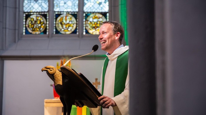 Ein Priester steht an einem Pult und spricht in eine Mikrofone, hinter ihm buntes Kirchenfenster.