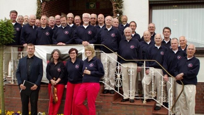 Ein großer Chor posiert vor einem Haus, einige Mitglieder stehen auf einer Treppe, andere vor einem Blumenbeet.