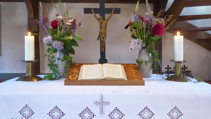 Eleganter Altar mit einem Kruzifix, einem aufgeschlagenen Buch, Kerzen und üppigen Blumenarrangements auf einem weißen, bestickten Tuch.