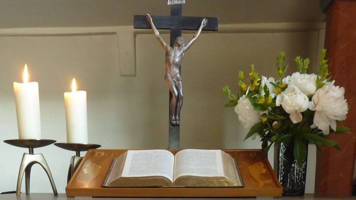 Eleganter Kirchenaltar mit Kerzen, einem Kreuz, einem aufgeschlagenen Buch, Blumen und einem mit Kreuz bestickten Leinentuch.