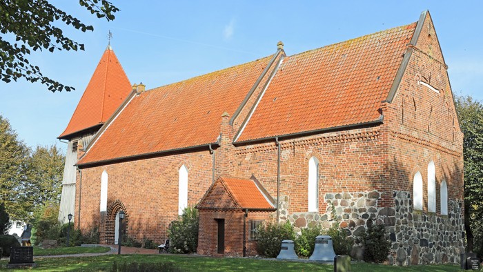 Das Bild zeigt eine rote Backsteinkirche mit spitzen Dächern und weißen Fenstern, umgeben von grünem Gras und Bäumen.