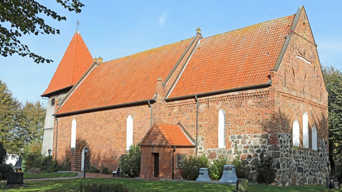 Das Bild zeigt eine rote Backsteinkirche mit spitzen Dächern und weißen Fenstern, umgeben von grünem Gras und Bäumen.