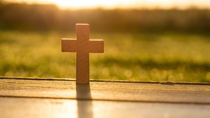 Une petite croix en bois posée sur une surface en bois devant un champ herbeux au coucher du soleil.