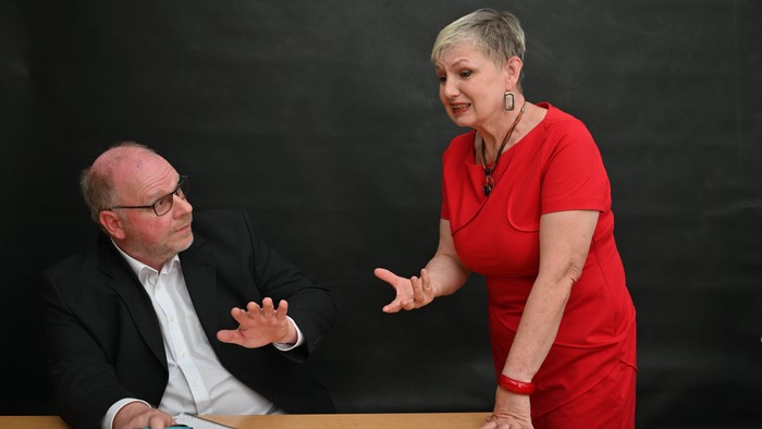 Zwei Personen im Gespräch, ein Mann im Anzug gestikuliert, während eine Frau in Rot lebhaft spricht.