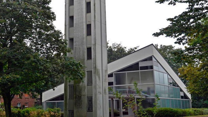 Das Bild zeigt eine moderne Kirche mit hohem Turm und Glasfront, umgeben von Bäumen.