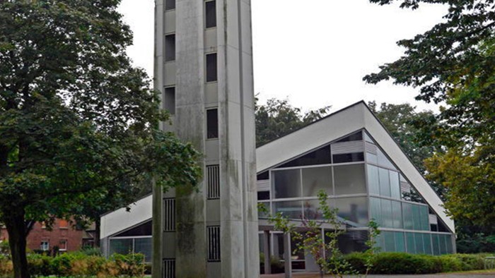 Das Bild zeigt eine moderne Kirche mit hohem Turm und Glasfront, umgeben von Bäumen.