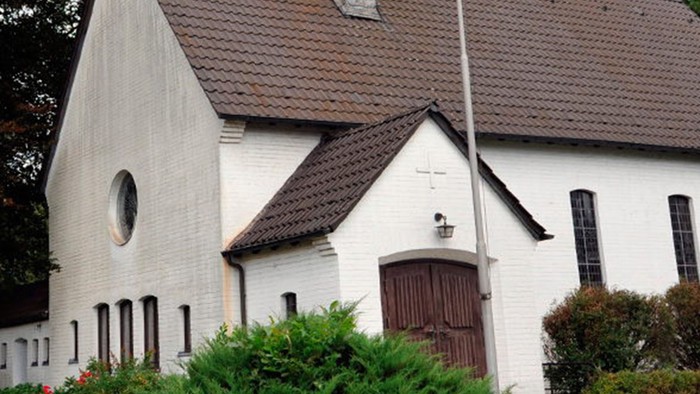 Weiße Kirche mit hohem Turm und braunem Dach, umgeben von Bäumen und Büschen