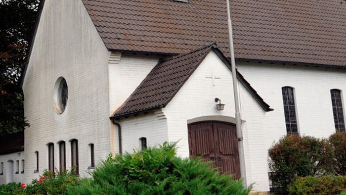 Weiße Kirche mit hohem Turm und braunem Dach, umgeben von Bäumen und Büschen