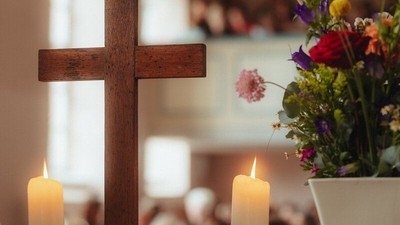 Ein ruhiger Altar zeigt ein hölzernes Kreuz mit brennenden Kerzen und eine Vase mit bunten Blumen in einer Kirchenumgebung.