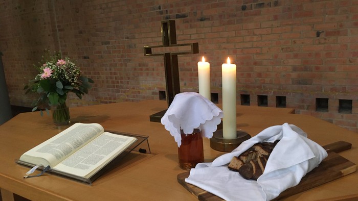 Religiöser Altaraufbau mit Kerzen, einem Kreuz, einem Buch, Blumen und einem bedeckten Laib Brot auf einem Holzbrett.